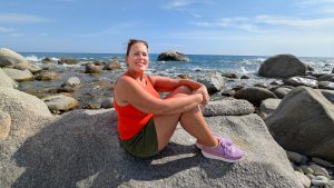 Kvinne smiler til kamera mens hun sitter i avslappet positur på en stor svaberglignende stein. Bakgrunnen er en steinstrand i Italia med mørkeblått hav og en lys, skyfri himmel.