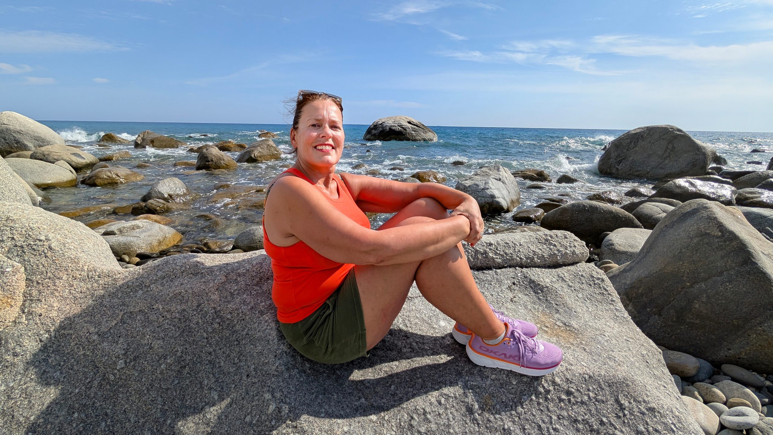 Kvinne smiler til kamera mens hun sitter i avslappet positur på en stor svaberglignende stein. Bakgrunnen er en steinstrand i Italia med mørkeblått hav og en lys, skyfri himmel.