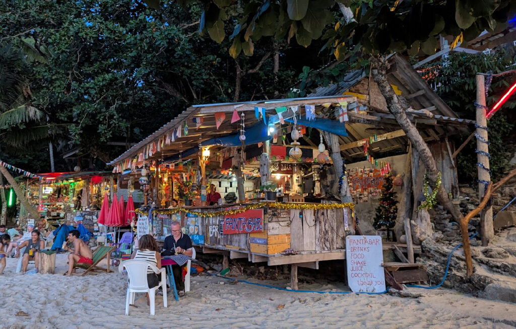 En rustikk strandbar bygget i tre på en sandstrand i skumringen. Baren er dekorert med fargerike flagg og lyslenker, og gjester sitter ved små bord i sanden foran baren