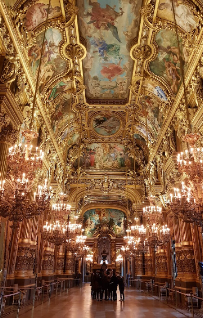 En enorm, overdådig hall i Opera Garnier med forgylte søyler, detaljerte takmalerier og rekker med store, lysende lysekroner. Gulvet har et sjakkbrettmønster i lyse farger, og folk står samlet i den fjerne enden av hallen.