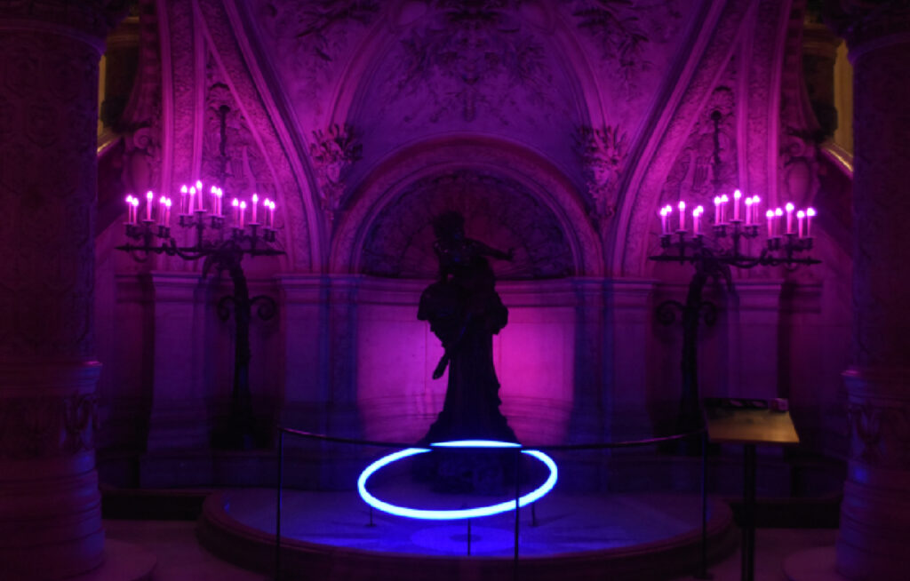 En klassisk statue i en buegang i kjelleren på Opera Garnier, opplyst av lilla kandelabre og en lysende blå sirkel på gulvet.