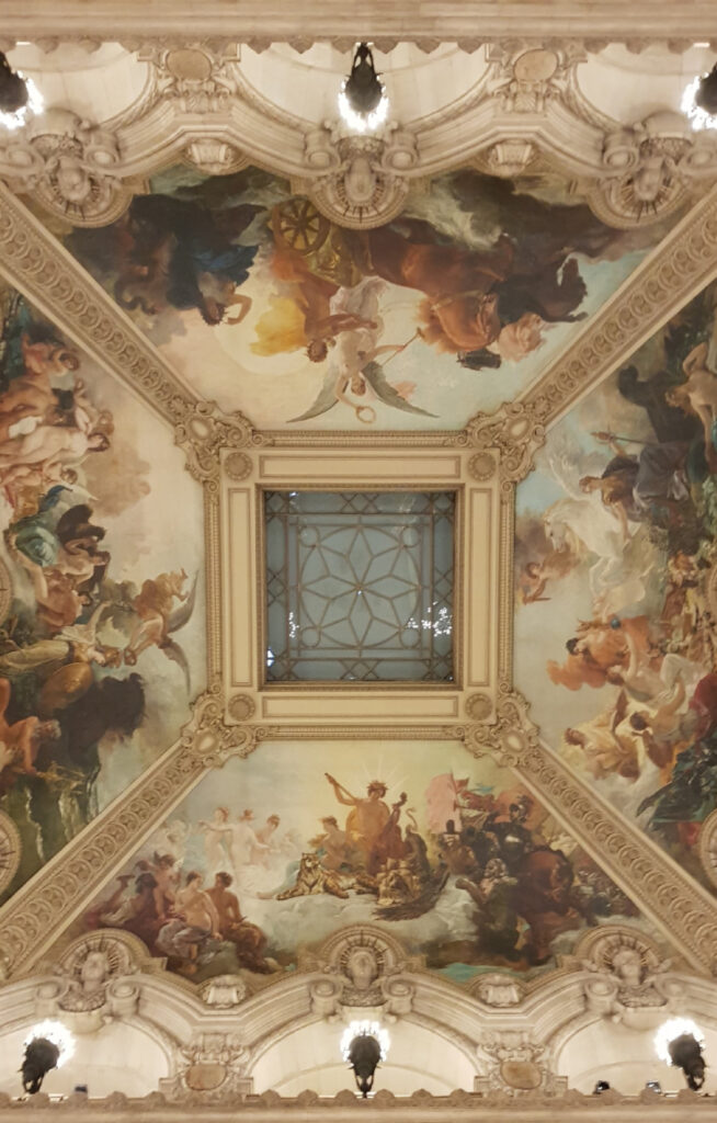 Et vertikalt bilde av den monumentale trappen i Opera Garnier. Man ser en marmorbalkong i midten, flankert av massive søyler og store kandelabre. Øverst i bildet vises det rikt dekorerte taket med malerier og stukkatur.