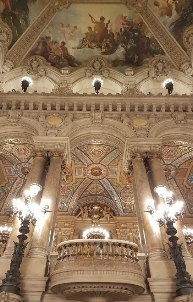 Et vertikalt bilde av den monumentale trappen i Opera Garnier. Man ser en marmorbalkong i midten, flankert av massive søyler og store kandelabre. Øverst i bildet vises det rikt dekorerte taket med malerier og stukkatur.