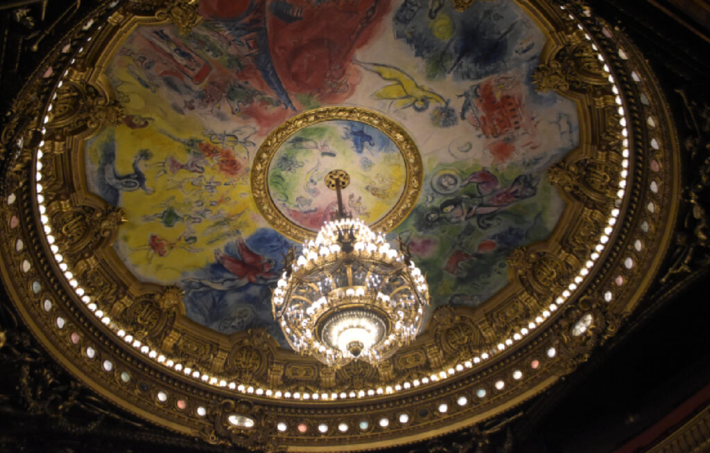 Et fargerikt, modernistisk takmaleri av Marc Chagall i Opera Garnier. En stor, glitrende krystallkrone henger i midten, omgitt av malte scener i gult, rødt, blått og grønt som forestiller kjente operaer og balletter.