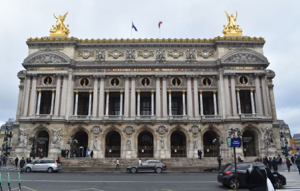 Fasaden til Palais Garnier i Paris under en overskyet himmel. Den monumentale bygningen har mange korintiske søyler, forgylte statuer på taket og sju store inngangsbuer på gateplan.