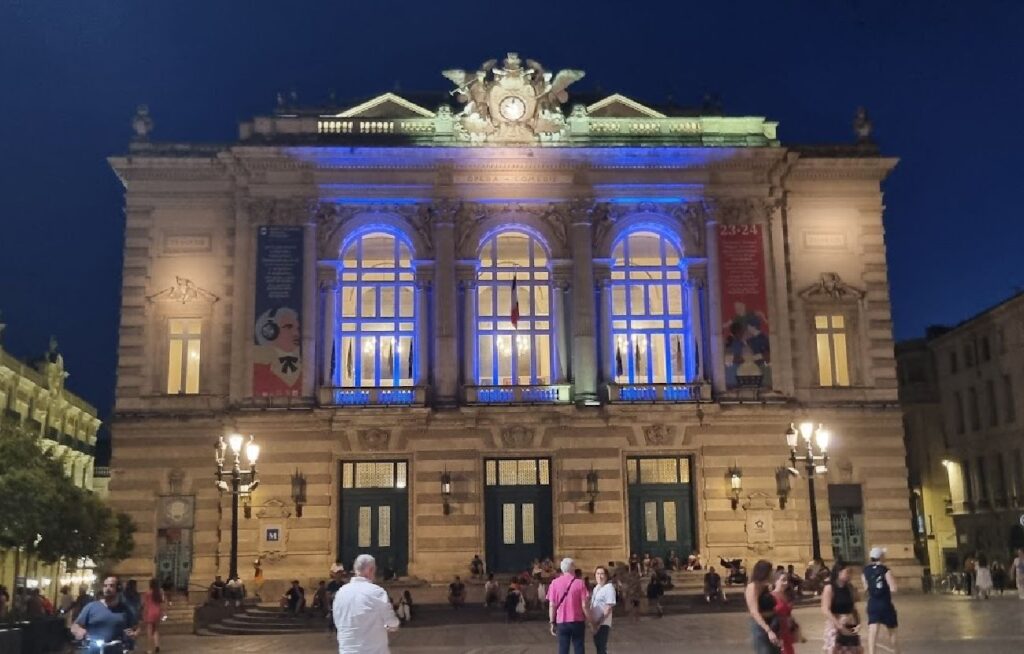 En stor, historisk operabygning i Montpellier fotografert om kvelden. Fasaden er vakkert belyst med gult og blått lys, og har store buevinduer og klassiske statuer på toppen. Mennesker oppholder seg på den åpne plassen foran bygningen.