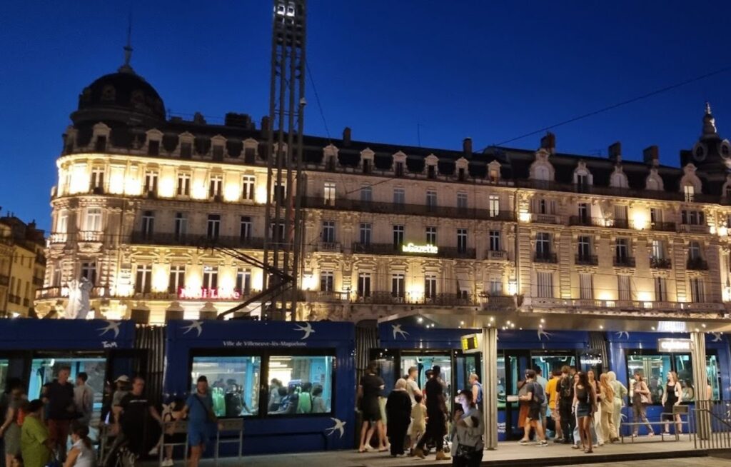En moderne blå trikk dekorert med hvite svaler står ved en holdeplass i Montpellier om kvelden. I bakgrunnen ses en staselig, opplyst historisk bygning med klassiske detaljer og franske balkonger mot en mørkeblå kveldshimmel.