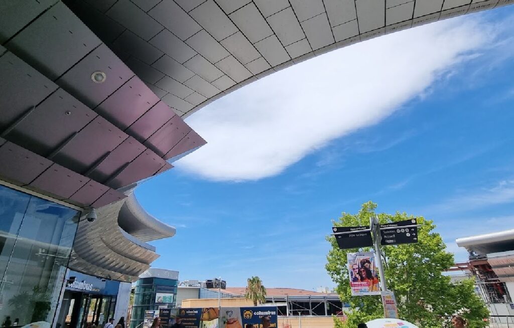 Moderne arkitektur med buede, metalliske takplater som rammer inn en blå himmel med en hvit sky. Under taket ser man inngangen til en kafé og et informasjonsskilt ved et utendørs kjøpesenter i Montpellier.