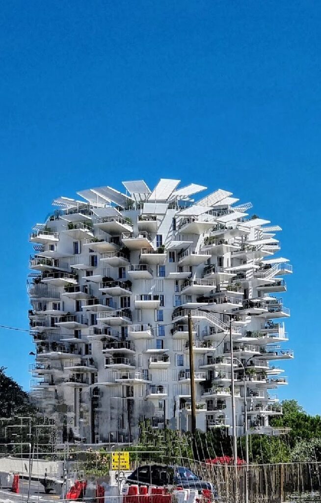 En unik hvit boligblokk med utallige tynne, utstikkende balkonger som minner om grener på et tre. Bygningen står mot en klar blå himmel i Montpellier, med grønne trær og en vei i forgrunnen.