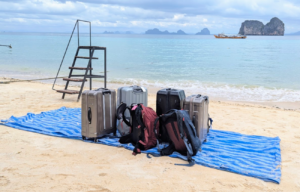En gruppe reisekofferter og ryggsekker står trygt samlet på en blå presenning på en hvit sandstrand med turkis hav og dramatiske klippeøyer i bakgrunnen.