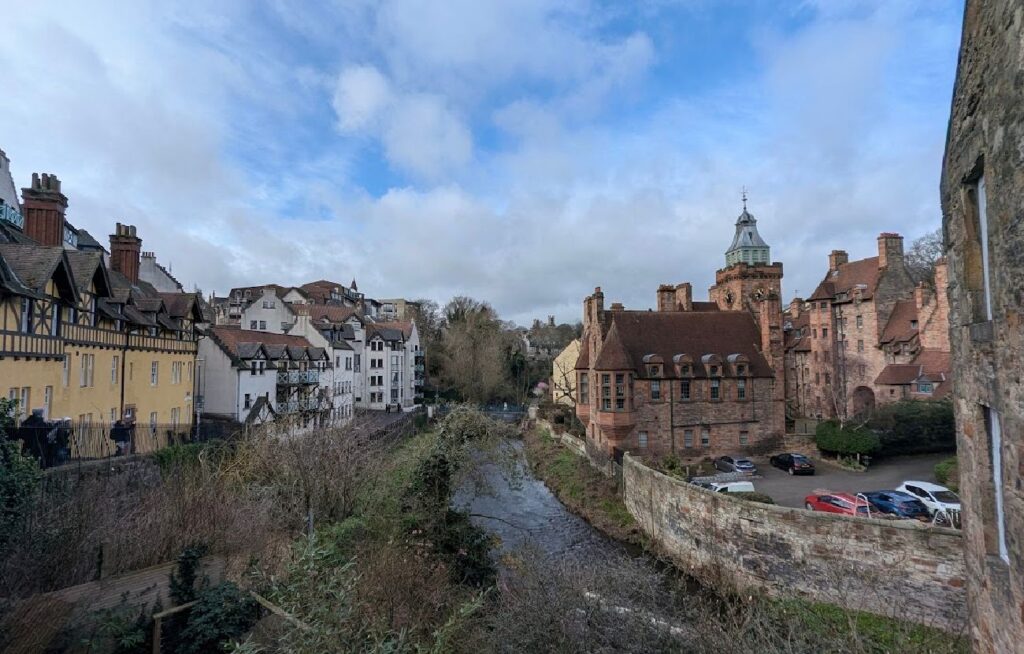 Historiske mursteinsbygninger med tårn og spir på venstre side av en elv, og en rekke sjarmerende gule hus på høyre side hvor turister går langs en gangvei i Dean Village.