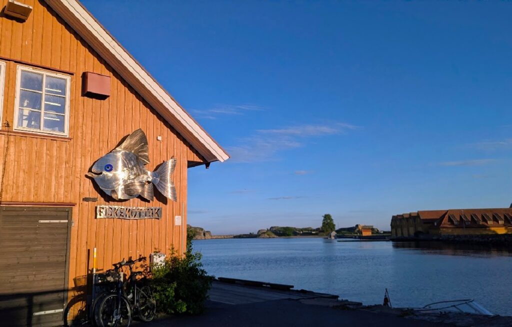 Det oransje trebygget til Stavern Fiskemottak på Dampskipsbrygga i Stavern, pyntet med en stor metallfisk på veggen, med blikkstille sjø og historiske bygninger i bakgrunnen.