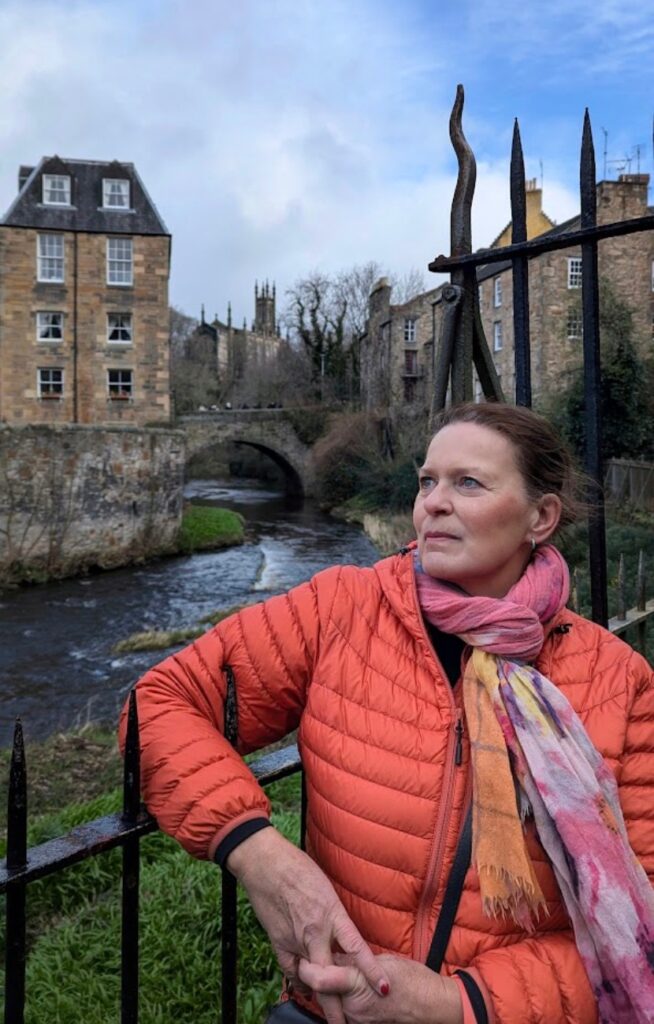 En kvinne i oransje jakke står ved et smijernsgjerde foran elven Water of Leith i Dean Village, Edinburgh, med historiske steinhus og en bro i bakgrunnen.