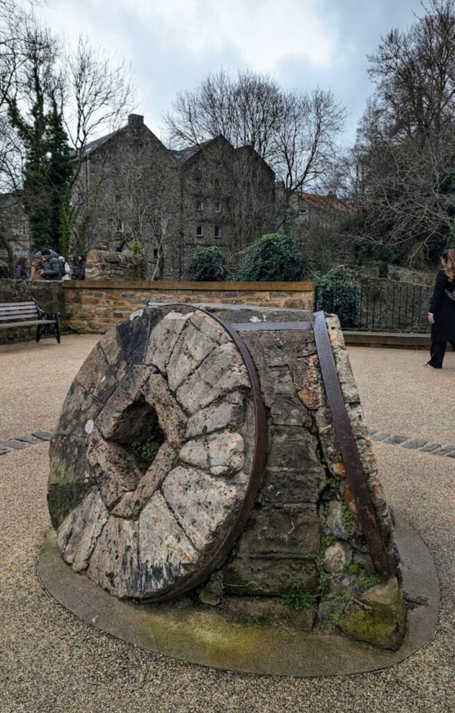 To store, gamle møllesteiner i stein og jern utstilt på en åpen plass i Dean Village, med historiske steinbygninger og nakne trær i bakgrunnen.