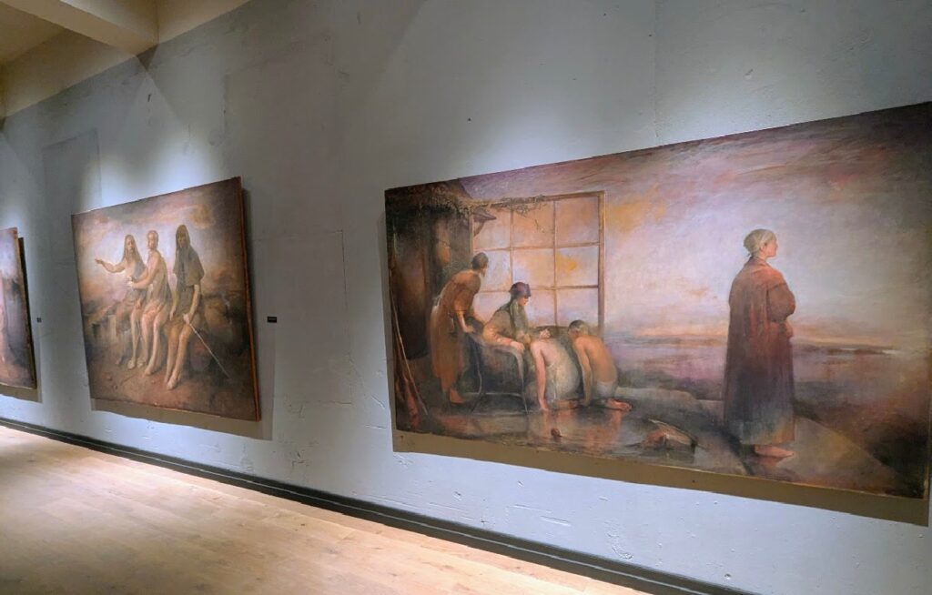 Monumentale oljemalerier av Odd Nerdrum utstilt langs en lang, grå gallerivegg med målrettet spotlight-belysning på Nerdrummuseet.