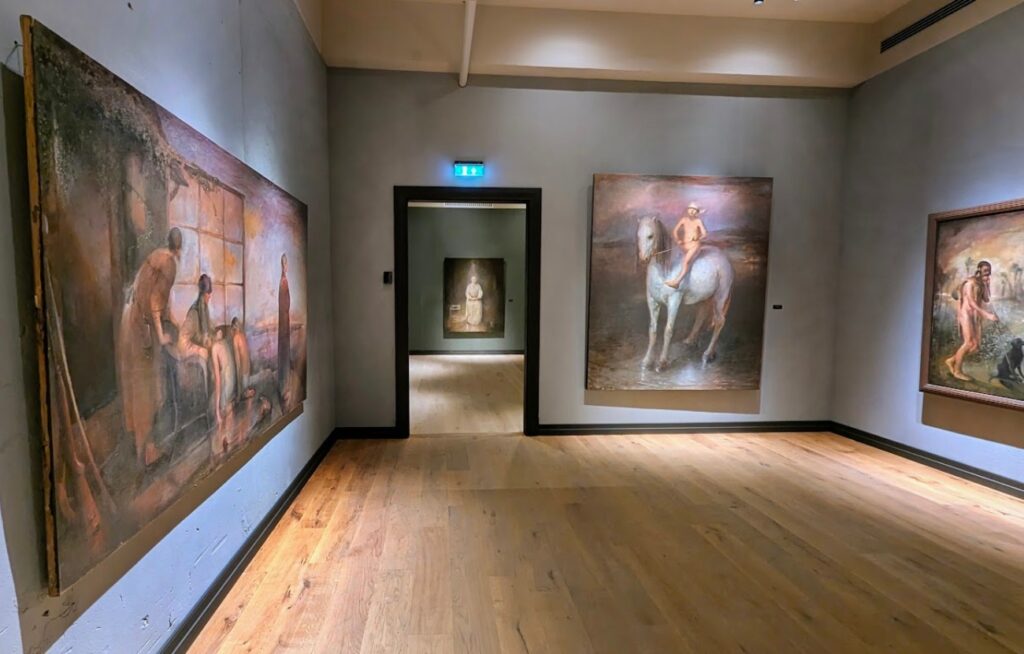 Stor utstillingssal på Nerdrummuseet med tregulv og grå vegger, som viser flere store kitch-malerier av Odd Nerdrum, inkludert et sentralt motiv av en person på en hvit hest.