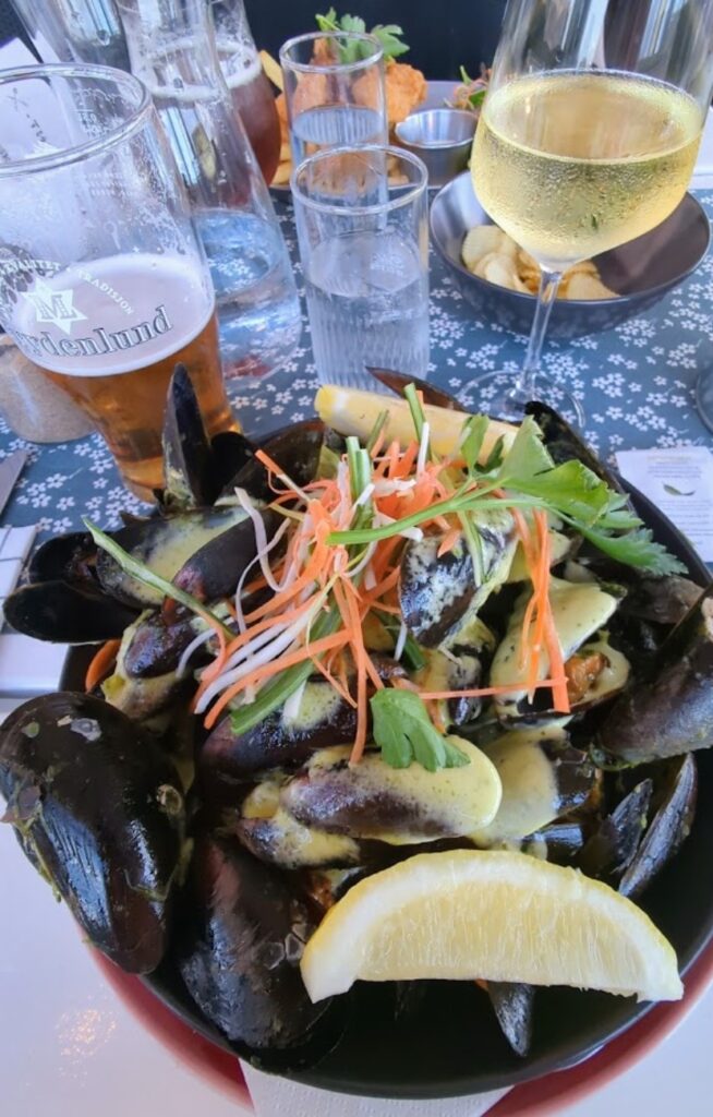 En dyp tallerken fylt med gratinerte blåskjell, urter, strimlede gulrøtter og en sitronbåt, servert på et restaurantbord med et glass hvitvin og et glass Frydenlund øl i bakgrunnen.
