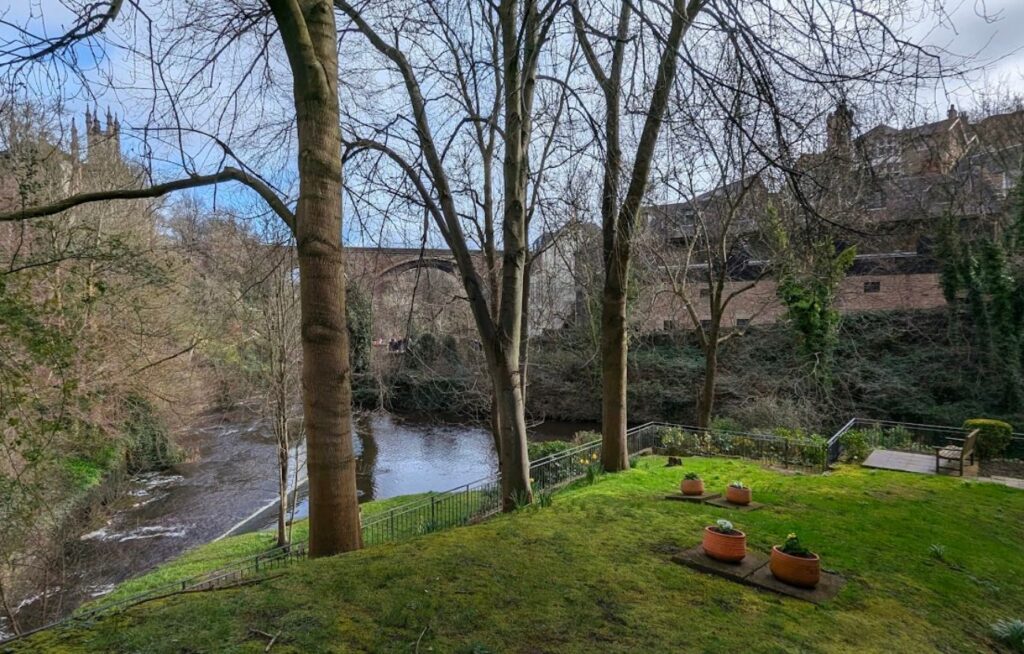 En rolig elv som renner gjennom en dal med grønne plener i forgrunnen, store trær uten blader, og en massiv steinbro (Dean Bridge) i bakgrunnen under en overskyet himmel i Edinburgh.
