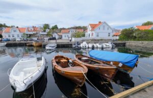 Trebåter og moderne plastbåter som ligger fortøyd i det blikkstille vannet i Nevlunghavn, med tradisjonelle hvite trehus i bakgrunnen under en lett skyet himmel.