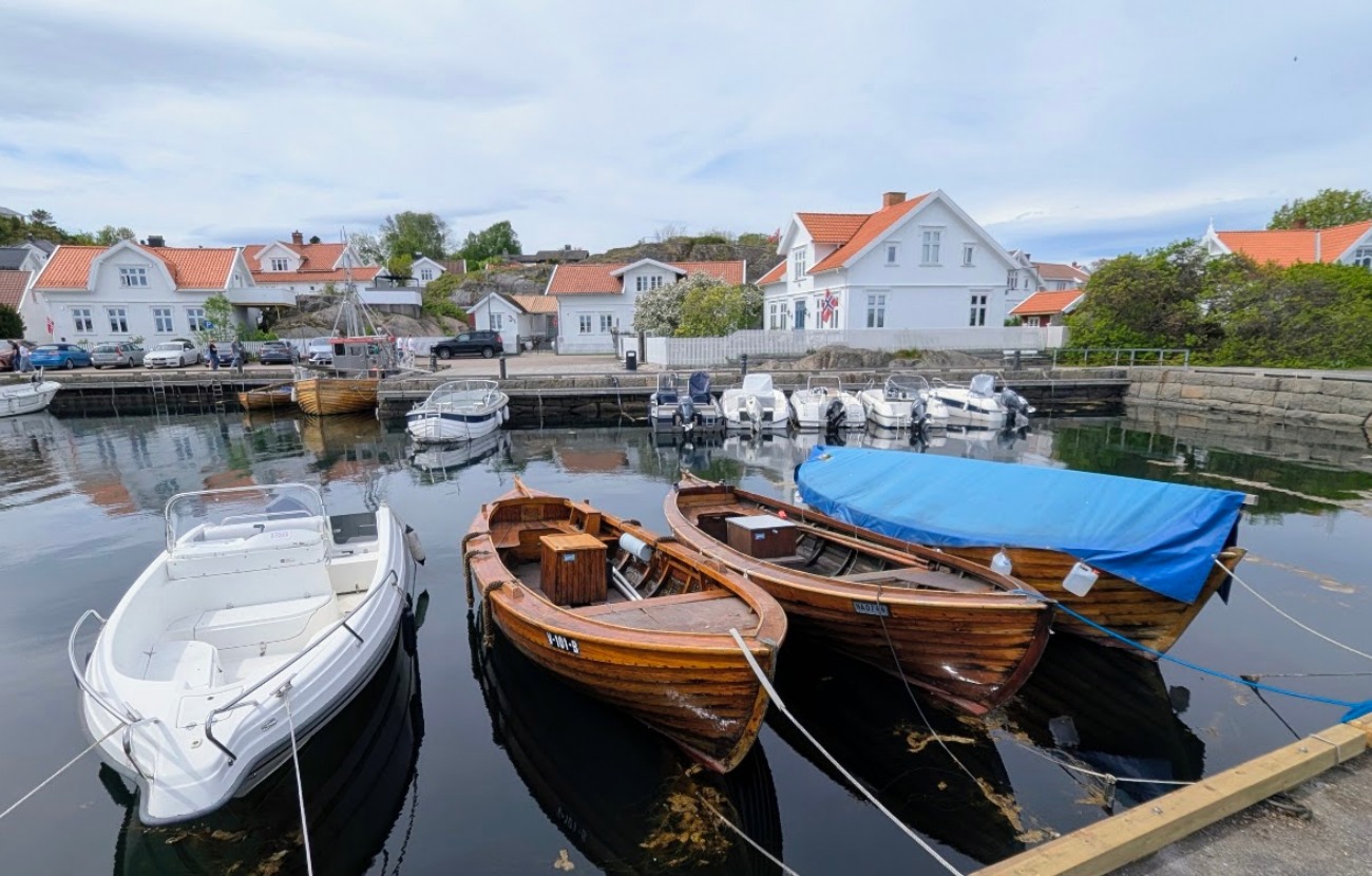 Trebåter og moderne plastbåter som ligger fortøyd i det blikkstille vannet i Nevlunghavn, med tradisjonelle hvite trehus i bakgrunnen under en lett skyet himmel.