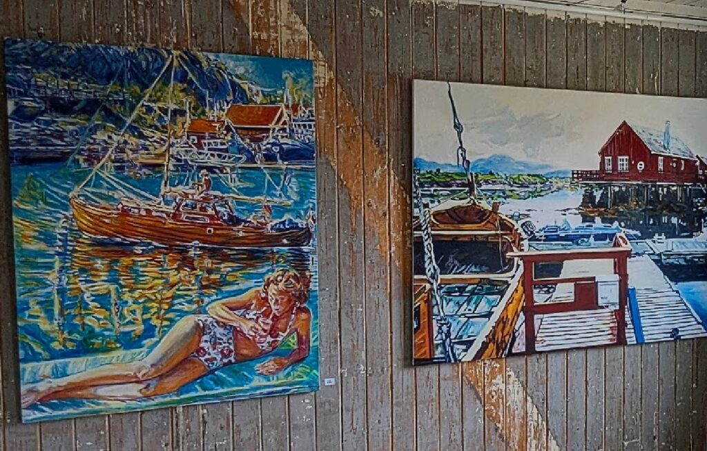 To store malerier med maritime motiver – ett av en kvinne på en flåte ved en seilbåt, og ett av en trebåt ved en sjøbod – henger på en slitt trevegg.