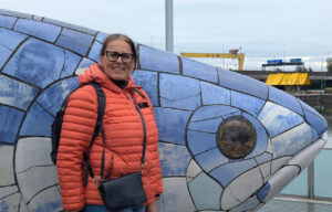 En smilende kvinne med briller og oransje dunjakke står foran den kjente keramikkskulpturen The Big Fish i Belfast.