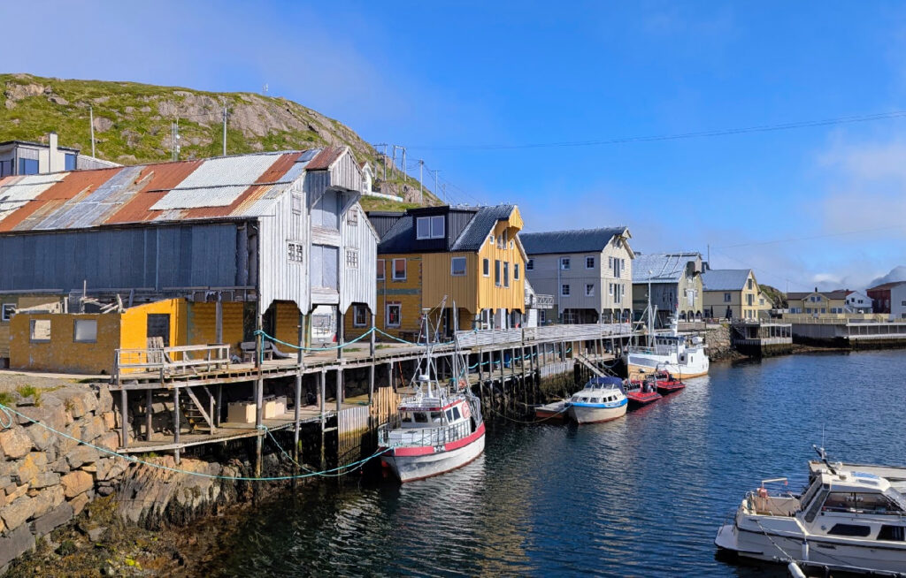 Oversiktsbilde av havnebassenget i Nyksund i Vesterålen, med fargerike sjøhus, brygger og flere fritidsbåter fortøyd langs kaien under en blå himmel.
