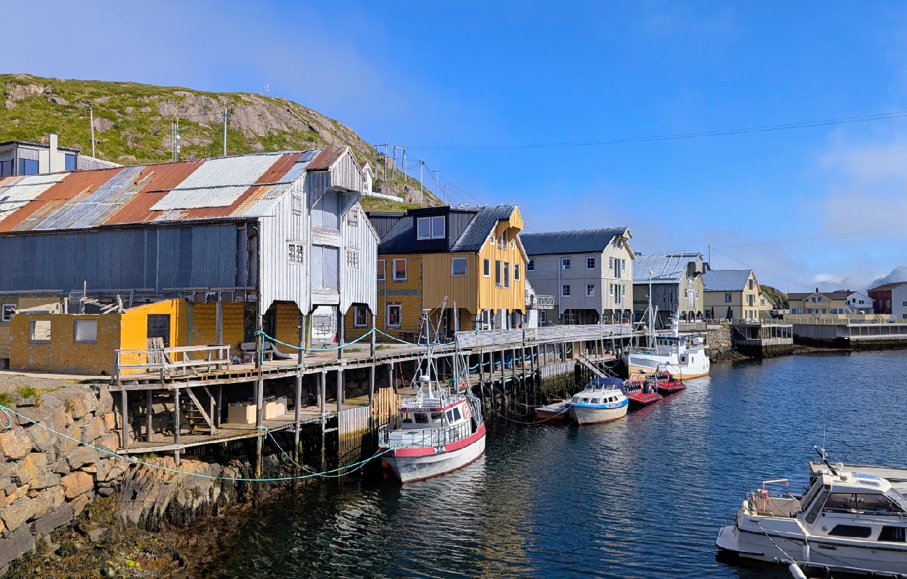 Oversiktsbilde av havnebassenget i Nyksund i Vesterålen, med fargerike sjøhus, brygger og flere fritidsbåter fortøyd langs kaien under en blå himmel.