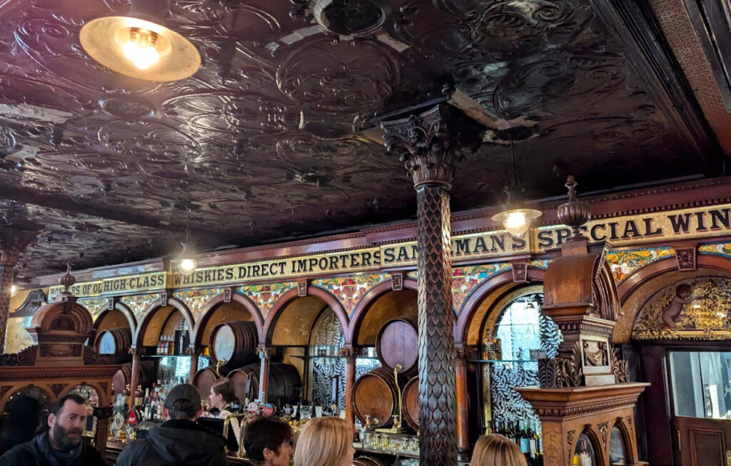 Interiørbilde av en tradisjonell og rikt dekorert bar med mørkt utskåret treverk, tak i messing med relieffer, og synlige øltønner bak bardisken.