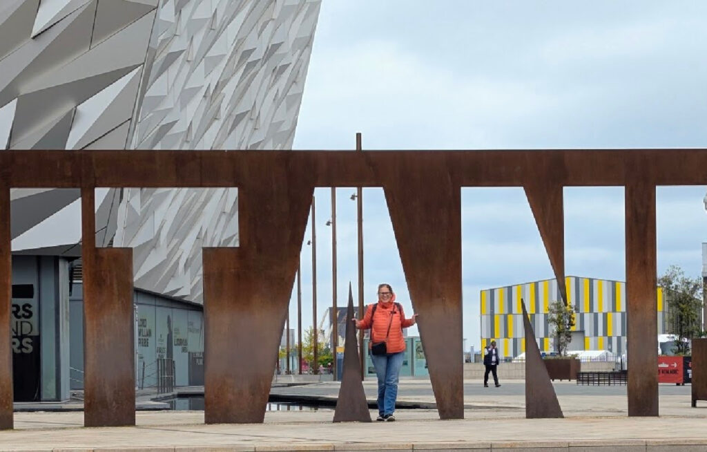 En kvinne i oransje jakke står i åpningen av de store, rustne bokstavene som staver TITANIC foran museumsbygget i Belfast.
