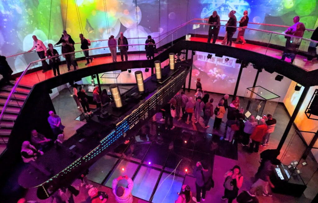 Oversiktsbilde fra innsiden av Titanic Belfast-museet med en stor skipsmodell i midten og besøkende i fargerikt lys.