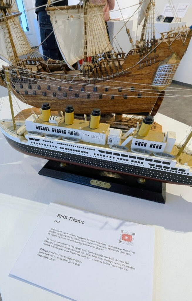 Nærbilde av en detaljert modell av RMS Titanic på en hvit utstillingsmonter, med et gammelt seilskip i bakgrunnen og en informasjonstavle i forgrunnen.