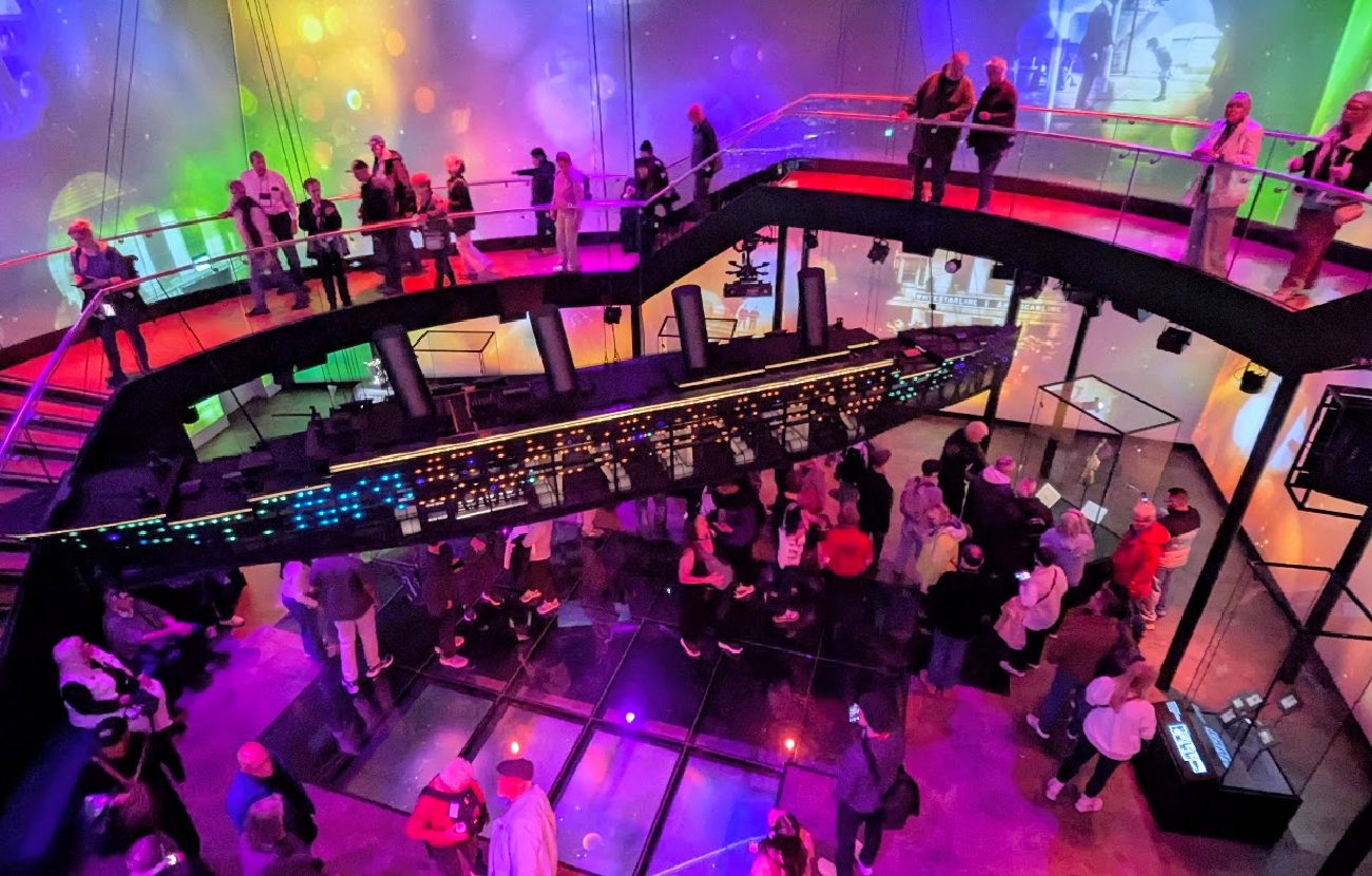 Besøkende som studerer utstillingen og skipsmodellen under fargerike projeksjoner inne på Titanic Belfast.