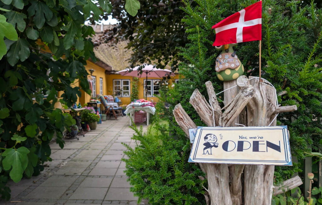 Inngangen til Anns lækkerier mikrobakeri i Lynæs, med et "Yes, we're OPEN"-skilt og et dansk flagg som ønsker gjester velkommen. En asfaltert sti fører gjennom en frodig hage til et gult hus med stråtak.