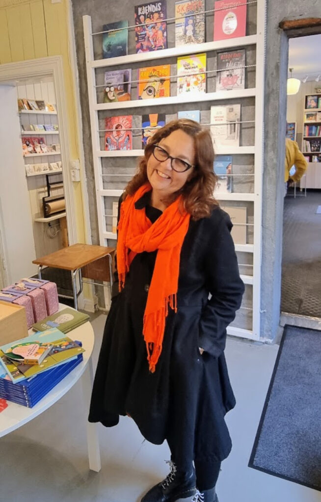 Smilende kvinne i svart kåpe og oransje skjerf blant bokhyller.