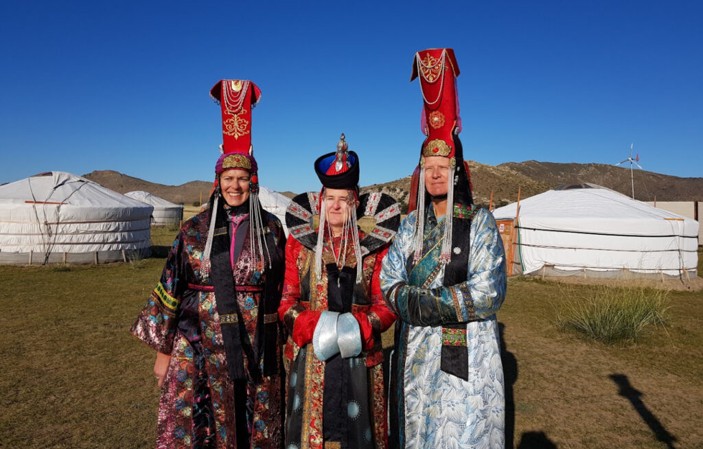 Tre vestlige reisende (to kvinner, en mann) står smilende side om side foran flere tradisjonelle hvite mongolske jurter. De er kledd i detaljerte, tradisjonelle mongolske drakter med fargerike kapper og forseggjorte hodeplagg. Bakgrunnen viser tørt gress, fjell og en klar blå himmel.