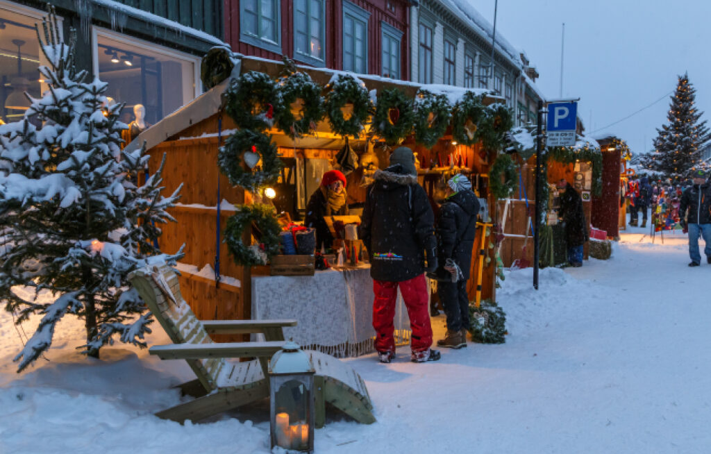 Snødekket julemarkedsbod på Røros med grankranser, varme lys, en trestol i forgrunnen og kledde besøkende i gaten om vinteren.