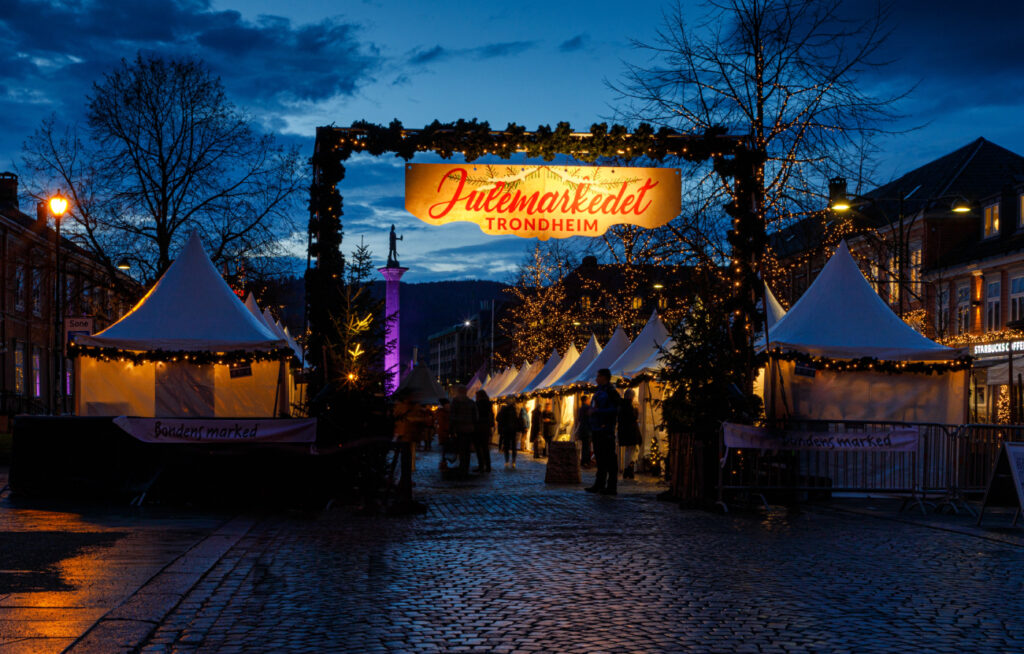 Den lysende portalen til Julemarkedet i Trondheim på Torvet i skumringen, med rekker av hvite teltboder og Olav Tryggvason-støtten i bakgrunnen.