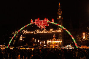 Den lysende inngangsportalen til Wiener Christkindlmarkt i Wien om kvelden, et av de mest populære julemarkeder i Europa, med folkemengder, karusell og det opplyste rådhustårnet i bakgrunnen.